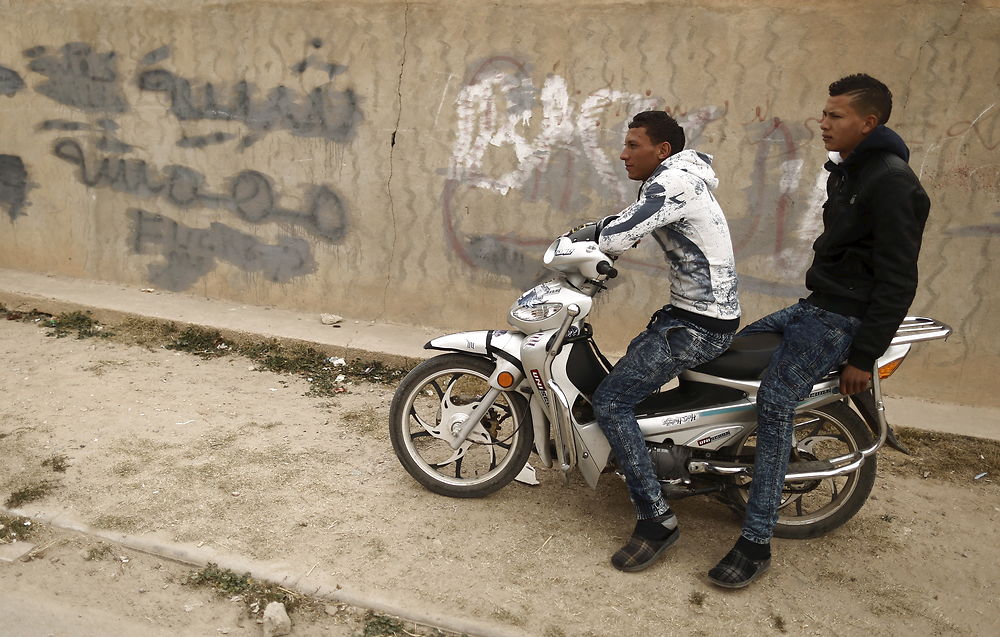 Arbejdsløse mænd på en motorcykel ved graffiti, hvor der står "Ungdommen er maginaliseret". I Zhor-kvarteret, Kasserine